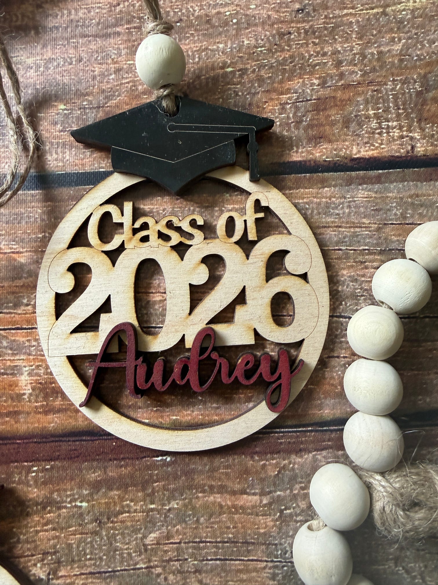 A personalized wooden graduation ornament featuring a black graduation cap and a round natural wood base with the text 'Class of 2026 Audrey' in light and dark red. The ornament is displayed on a rustic wooden surface next to a wooden bead garland.