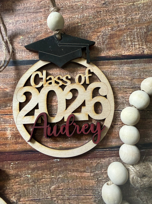 A personalized wooden graduation ornament featuring a black graduation cap and a round natural wood base with the text 'Class of 2026 Audrey' in light and dark red. The ornament is displayed on a rustic wooden surface next to a wooden bead garland.