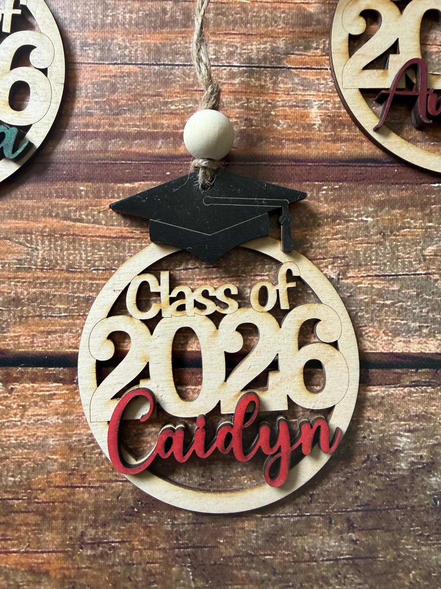 A wooden graduation ornament with a black mortarboard cap and a hanging string, featuring the cutout text 'Class of 2026' and the name 'Caidyn' in red, displayed on a rustic wood background.