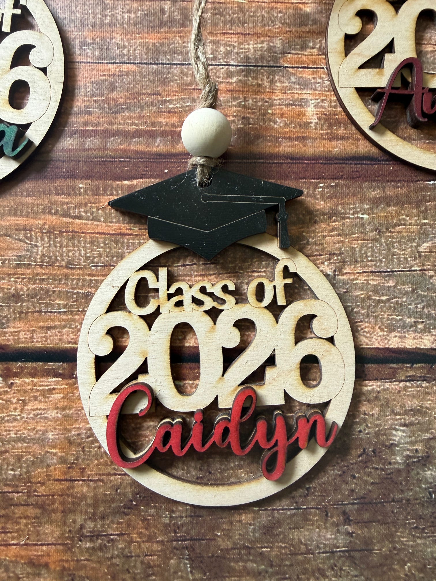 A wooden graduation ornament with a black mortarboard cap and a hanging string, featuring the cutout text 'Class of 2026' and the name 'Caidyn' in red, displayed on a rustic wood background.