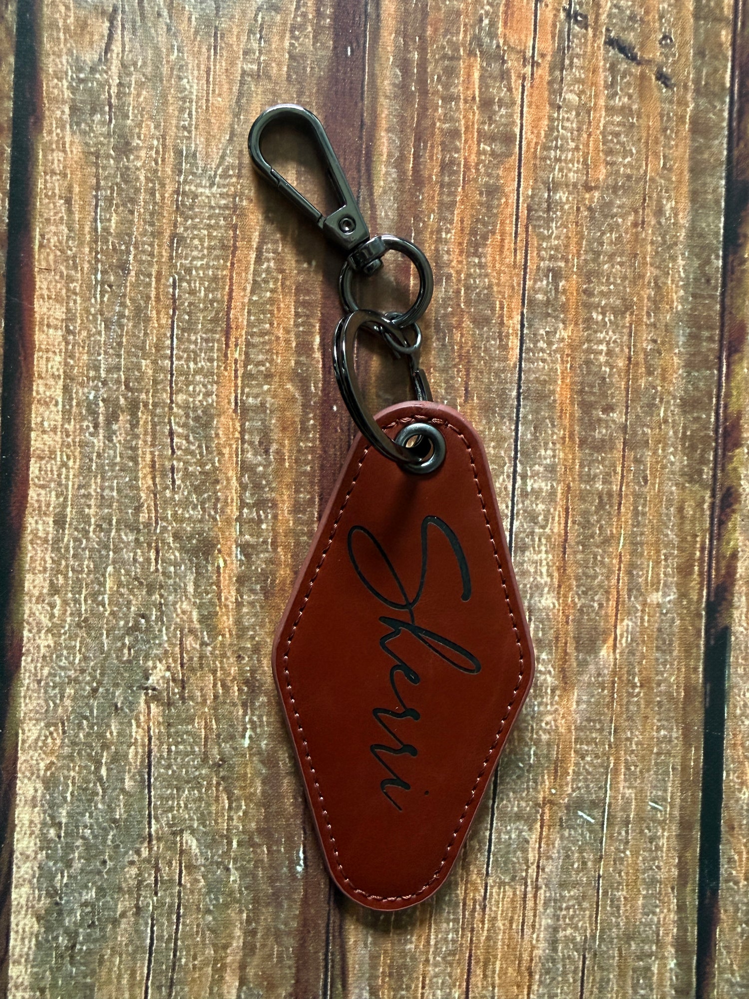 A leather keychain tag with the name written in black script, attached to a black metal swivel clasp, displayed on a rustic wooden surface.