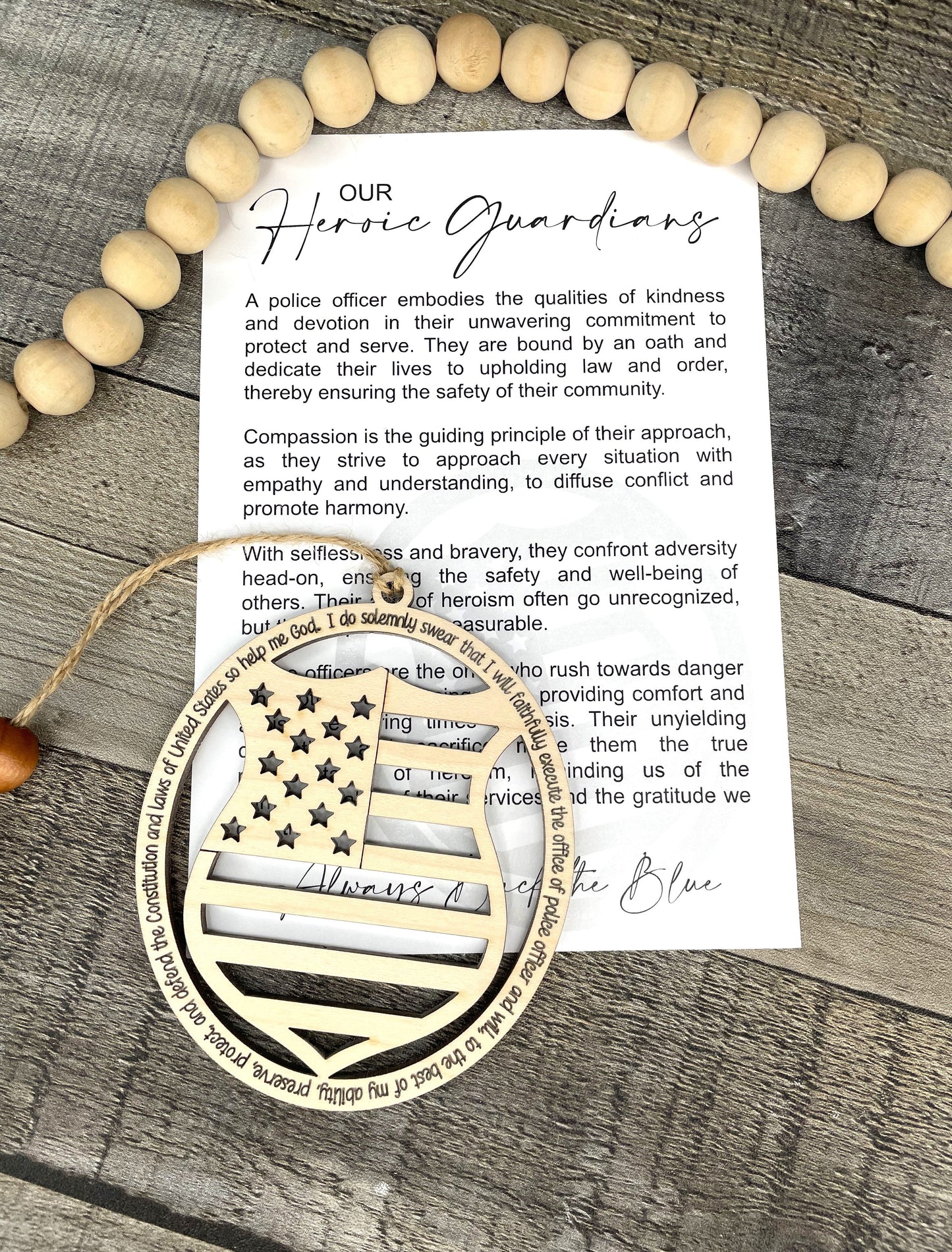 A printed card titled 'Our Heroic Guardians' with a quote about police officers, displayed on a rustic gray wood surface with wooden ornament that reflects a USA flag and badge. 