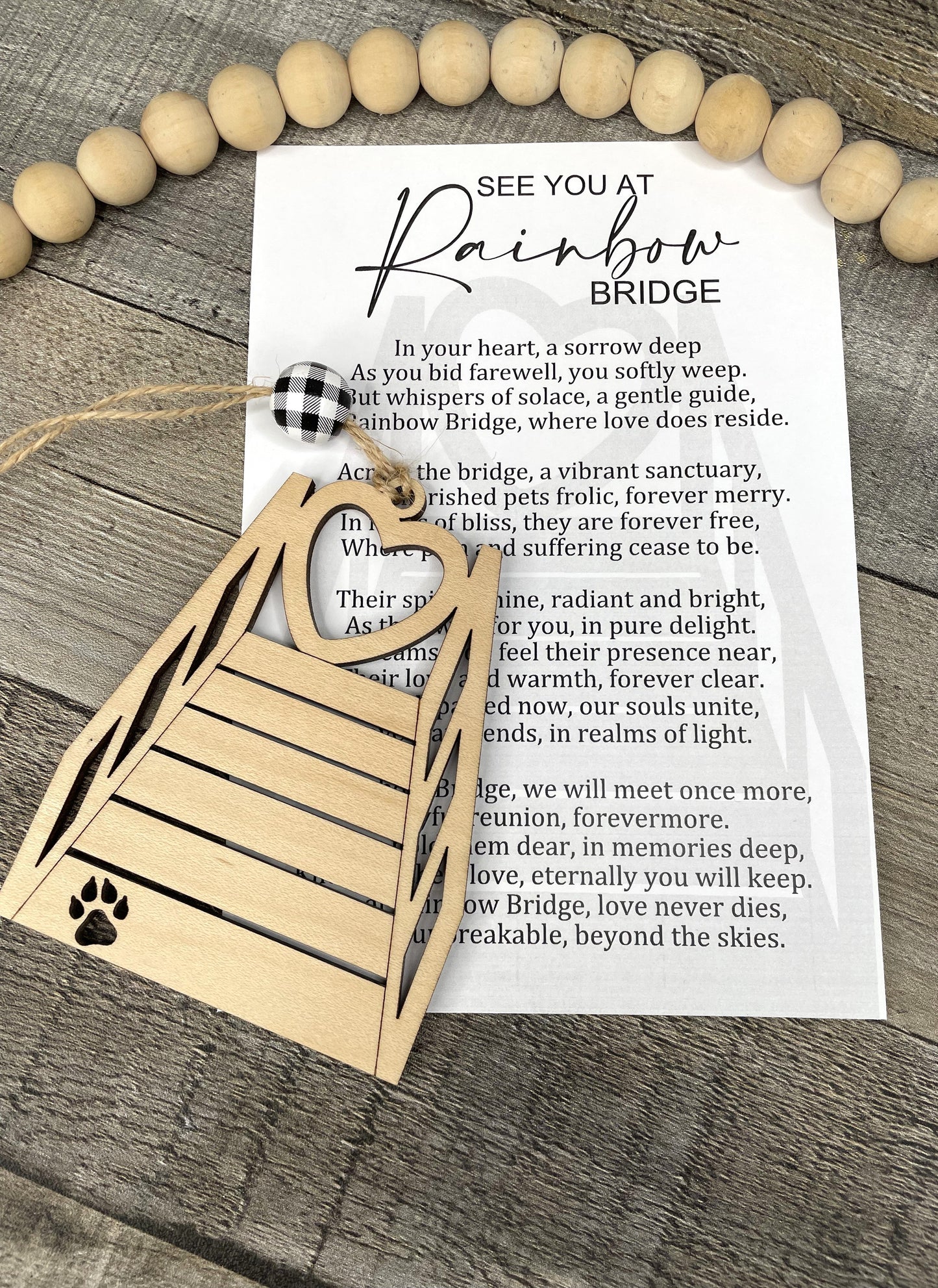 A wooden memorial ornament with a paw print and rainbow bridge design, displayed on a rustic wood background next to a printed 'See You At Rainbow Bridge' card.