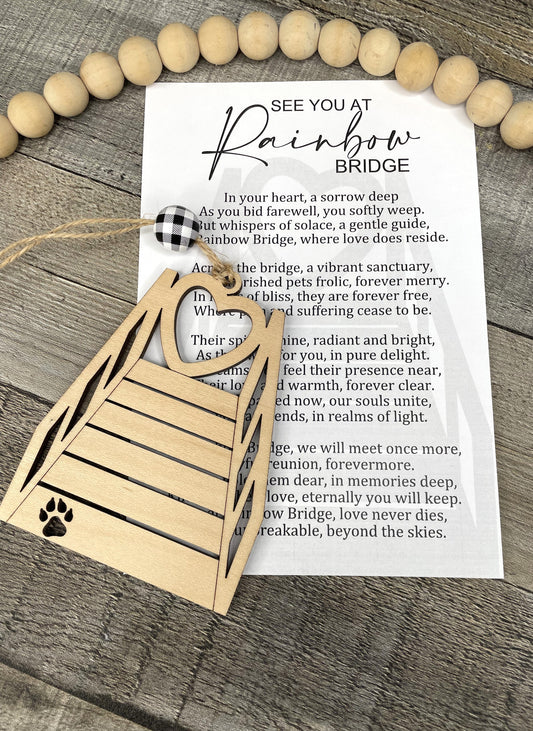 A wooden memorial ornament with a paw print and rainbow bridge design, displayed on a rustic wood background next to a printed 'See You At Rainbow Bridge' card.