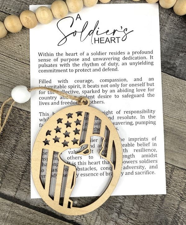 A wooden patriotic ornament with an American flag design, displayed on top of a printed card with the title 'A Soldier's Heart' on a rustic wood background.