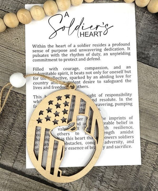 A wooden patriotic ornament with an American flag design, displayed on top of a printed card with the title 'A Soldier's Heart' on a rustic wood background.