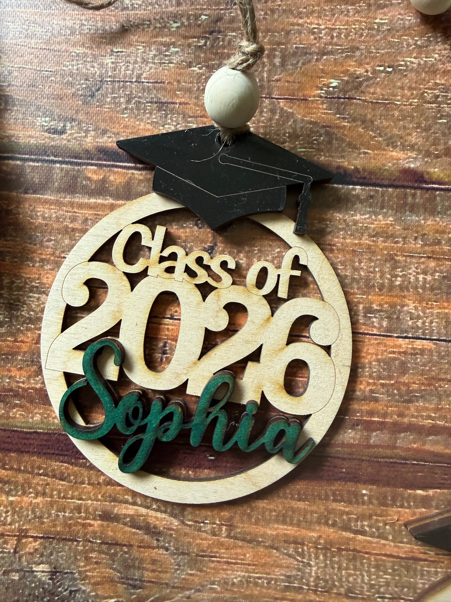 A wooden graduation ornament with a black graduation cap and a hanging bead, featuring laser-cut text reading 'Class of 2026' and 'Sophia' on a round light wood base, photographed on a rustic wooden surface.