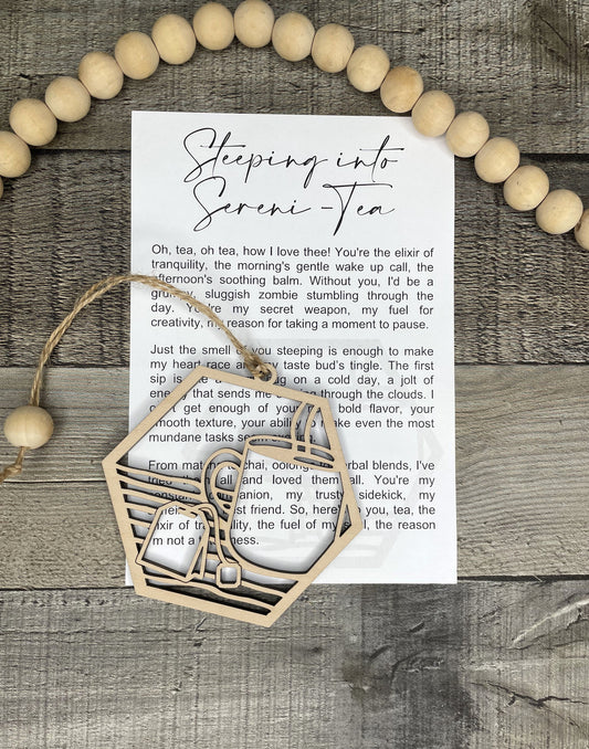 A wooden laser-cut tea cup and tea bag ornament lies on a printed poem card, surrounded by a wooden bead garland and set on a rustic gray wood surface.