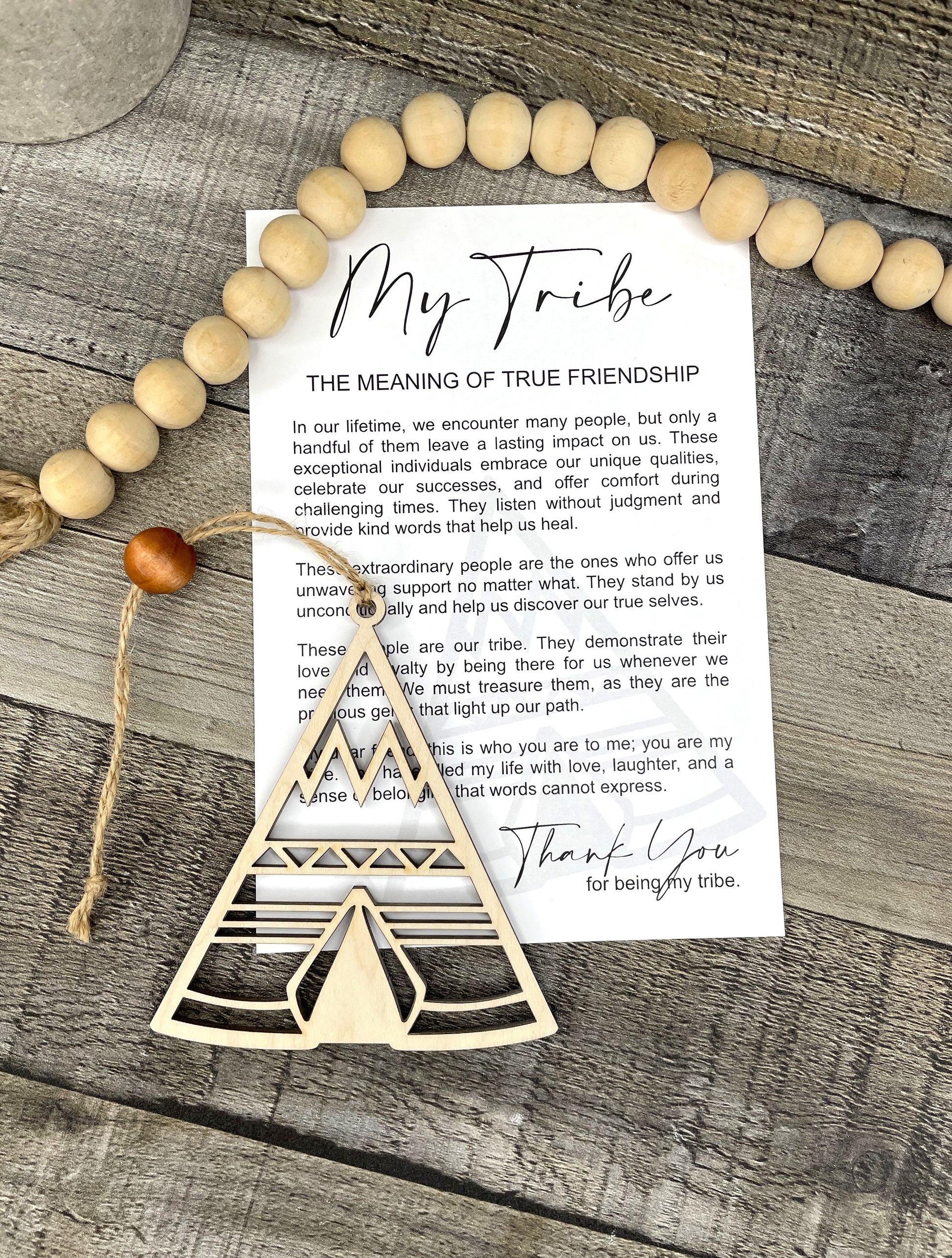 A wooden Tribe ornament and a printed card reading 'My Tribe: The Meaning of True Friendship' are arranged on a rustic wood surface with a beaded garland.