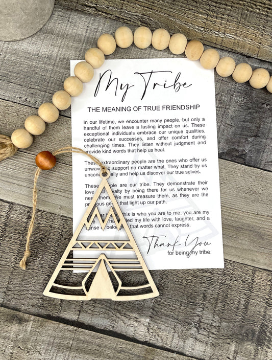 A wooden Tribe ornament and a printed card reading 'My Tribe: The Meaning of True Friendship' are arranged on a rustic wood surface with a beaded garland.